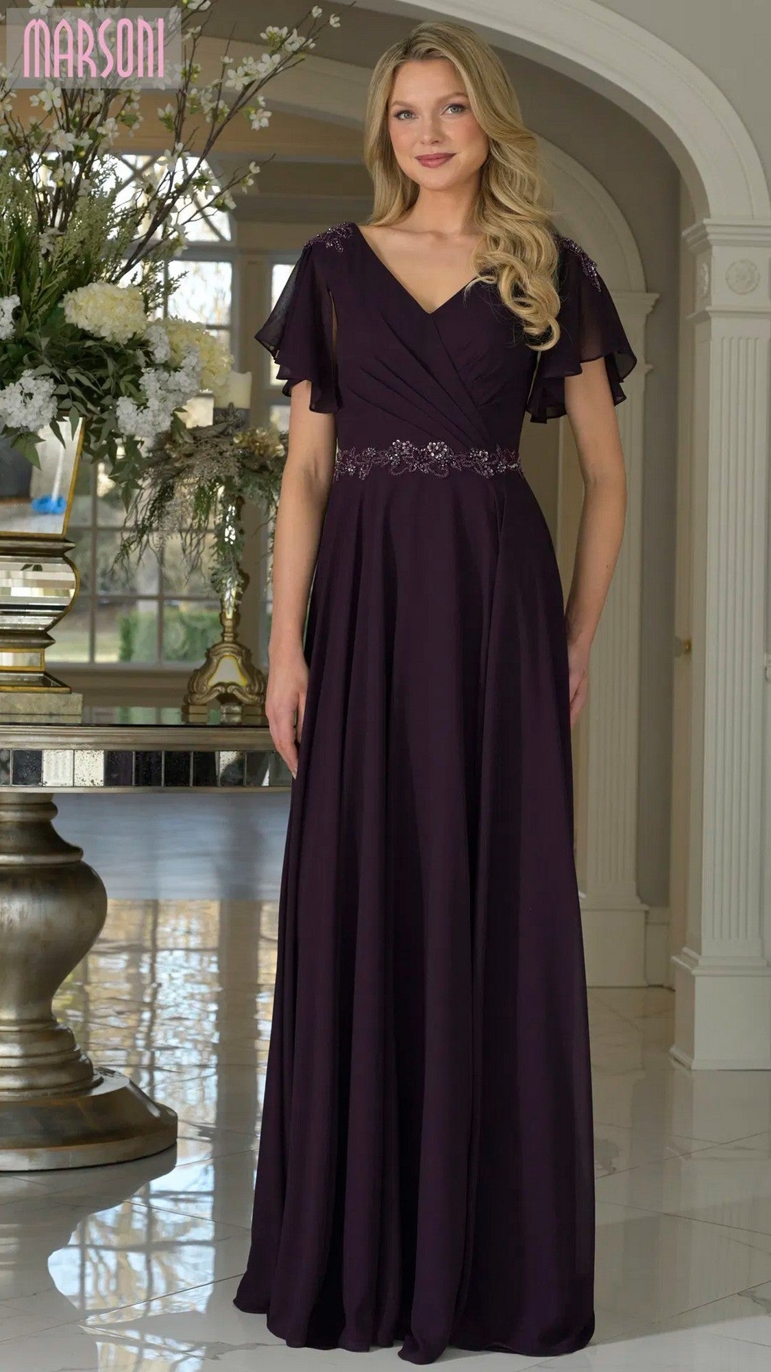 A woman stands indoors wearing the Marsoni M328 long formal dress in eggplant chiffon, featuring short flutter sleeves, a beaded embellished waistband, and an elegant a-line skirt.
